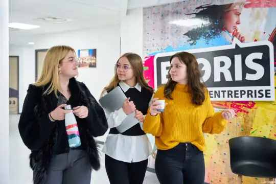 Trois étudiantes d'Ecoris discutent ensemble dans un couloir de l'école, devant un mur coloré avec le logo Ecoris.