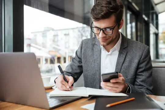 Jeune homme en costume gris travaillant sur son ordinateur portable dans un café, prenant des notes tout en consultant son smartphone