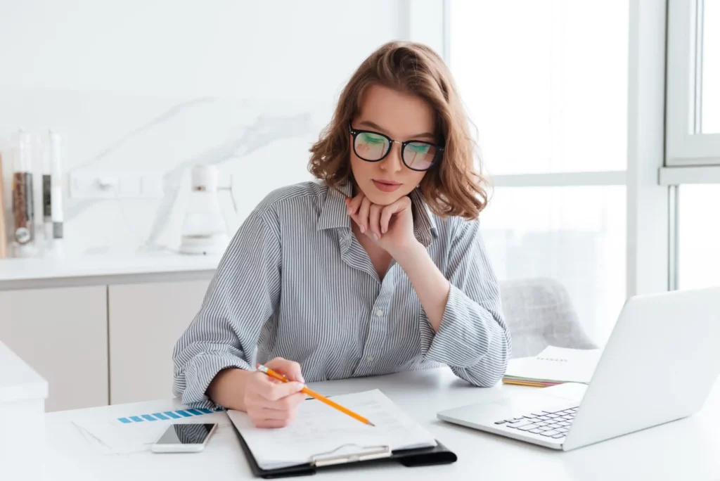 Jeune femme portant des lunettes et une chemise rayée, concentrée sur des documents à son bureau à domicile, dans le cadre d’une formation en alternance Ecoris en commerce et gestion.