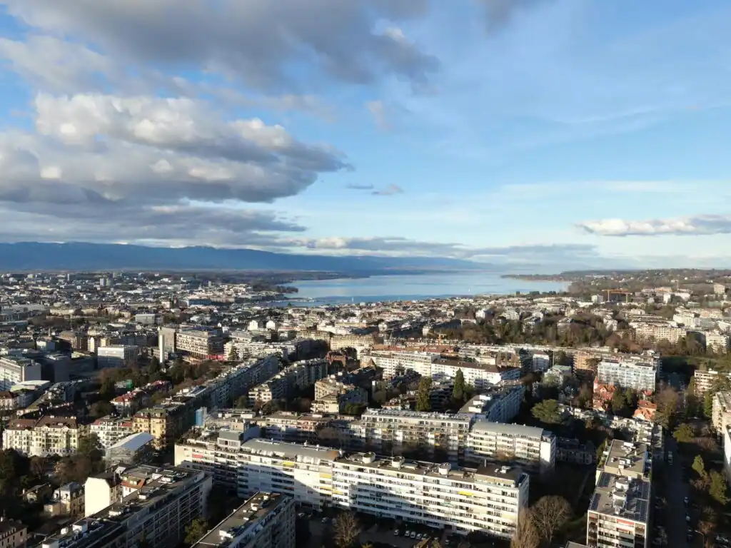 Vue aérienne de Genève avec le lac Léman en arrière-plan, sous un ciel partiellement nuageux.