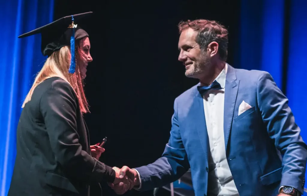 Une diplômée d'Ecoris Chambéry reçoit son diplôme sur scène, félicité par un représentant de l'école lors de la cérémonie officielle.
