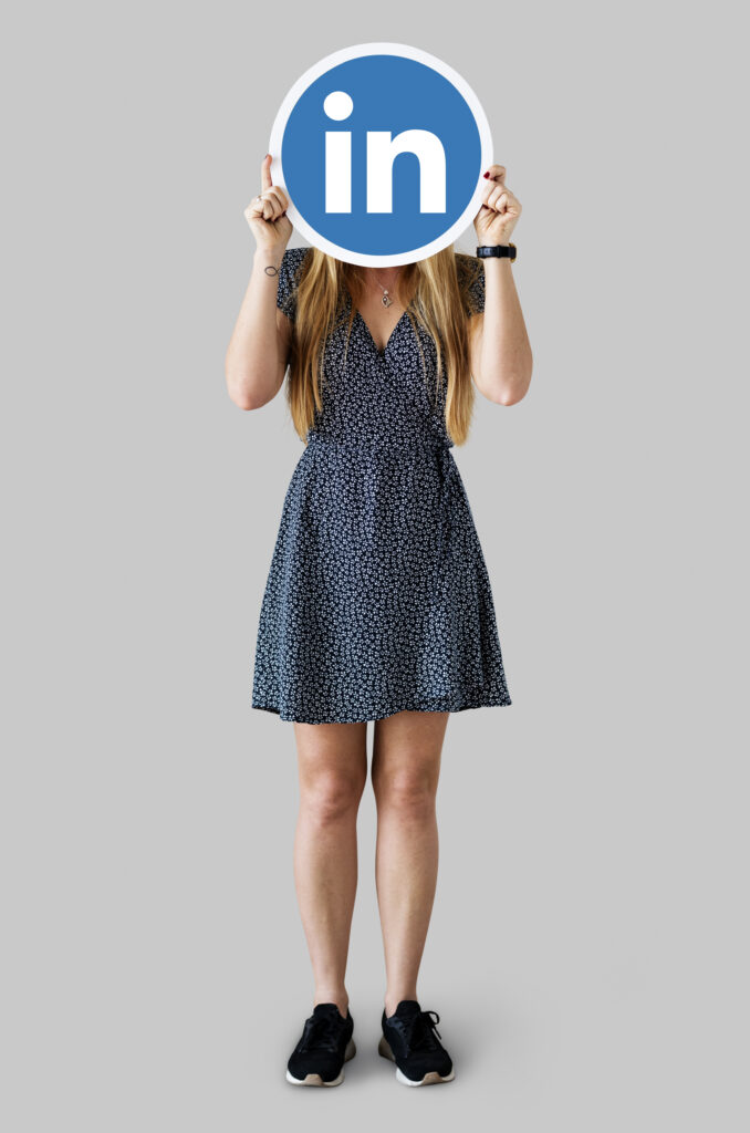 Femme en robe tenant un logo LinkedIn devant son visage sur fond gris, symbolisant la présence en ligne et le personal branding.